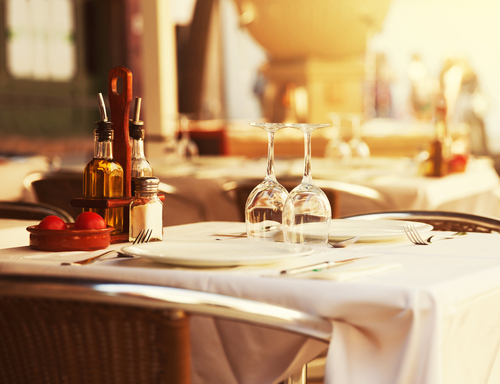 A restaurant table with white tablecloth, two upside-down wine glasses, a plate with cutlery, and a wooden caddy holding oil, vinegar, and salt beside a bowl of tomatoes.