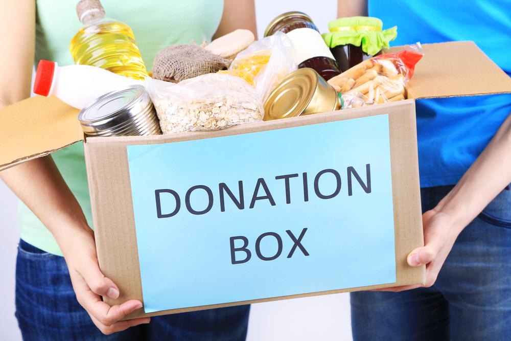  Cardboard box labeled "DONATION BOX" filled with various non-perishable food items, held by two people.