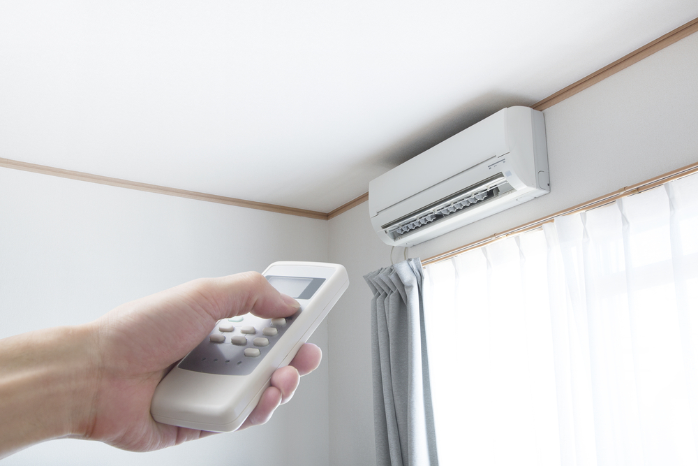  A hand holding a remote pointed at a wall-mounted air conditioner near a window with white curtains.