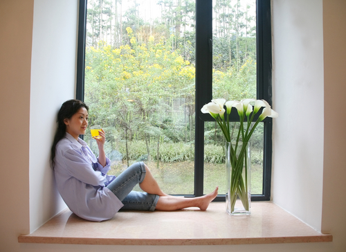 Woman sitting on a windowsill holding a yellow glass, with a vase of white flowers next to her and a view of trees and yellow flowers outside.