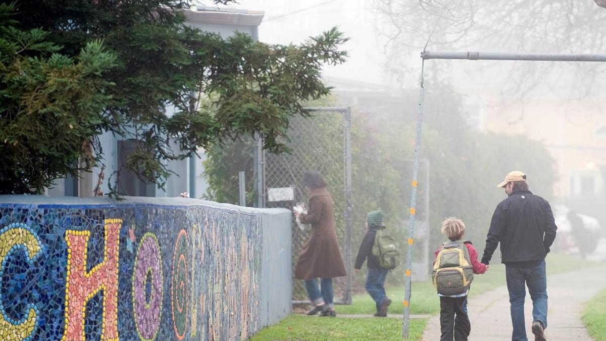 A foggy morning scene of an adult and a child walking past a colorful, mosaic-tiled school wall.
